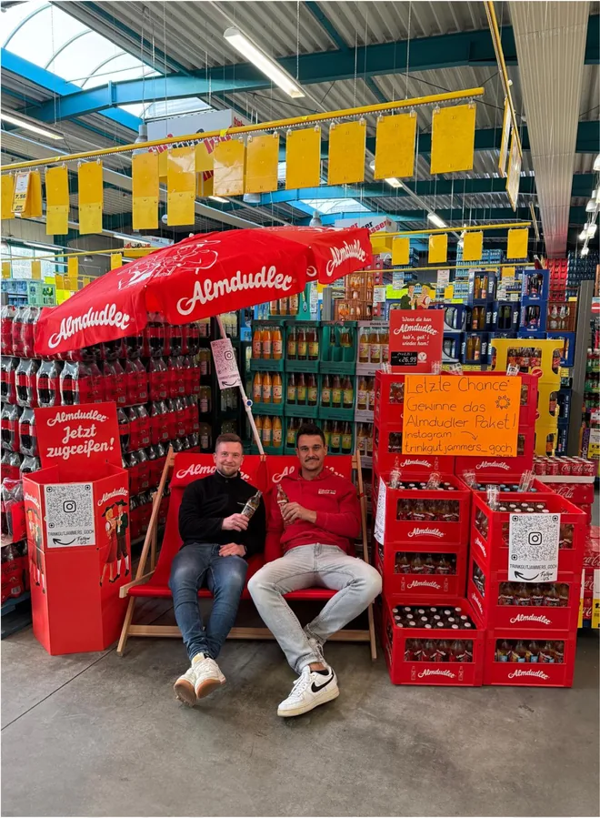 Twee personen op een bank onder een rode Almdudler-parasol tussen drankkratten